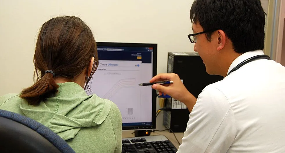 A clinician in a white coat points a pen at a chart on a desktop monitor while a patient, seen from behind, sits beside him in a small consulting room.