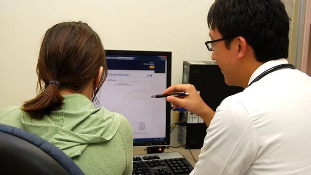 A clinician in a white coat points a pen at a chart on a desktop monitor while a patient, seen from behind, sits beside him in a small consulting room.