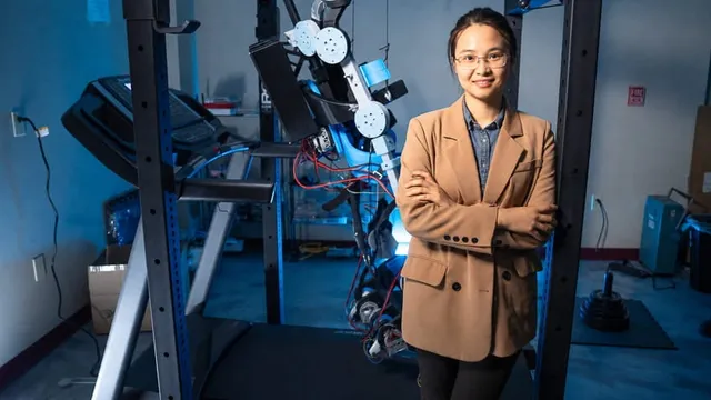 Shuzhen Luo stands with folded arms beside a wearable exoskeleton with exposed motors and wiring, mounted next to a treadmill in a blue-lit Embry-Riddle robotics laboratory.