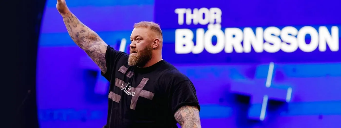 A heavily built bearded athlete in a black T-shirt reading I am Enhanced raises one tattooed arm on a blue-lit arena stage, his name on the screen behind him.