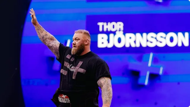A heavily built bearded athlete in a black T-shirt reading I am Enhanced raises one tattooed arm on a blue-lit arena stage, his name on the screen behind him.