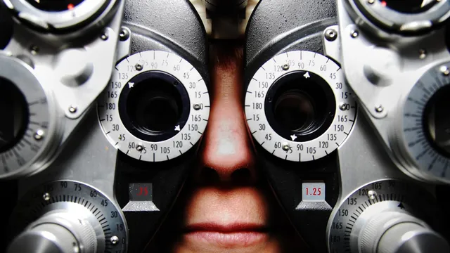 Close-up of a phoropter with two lens turrets and numbered dial rings, a patient nose and mouth visible behind the eyepieces during an eye exam.