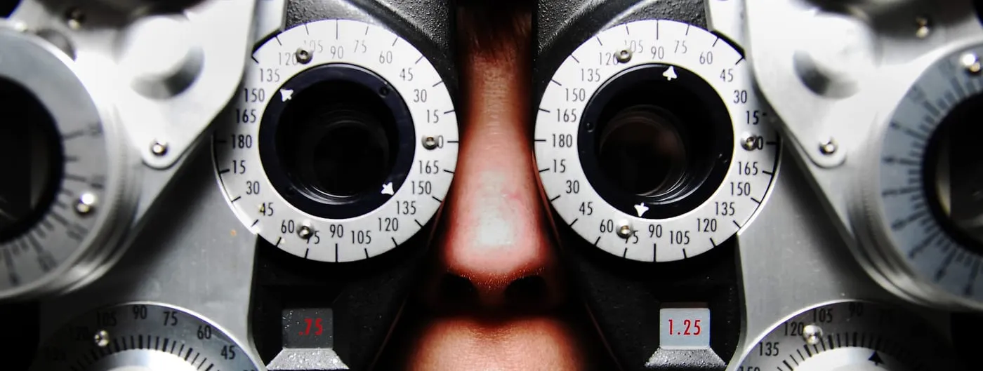 Close-up of a phoropter with two lens turrets and numbered dial rings, a patient nose and mouth visible behind the eyepieces during an eye exam.