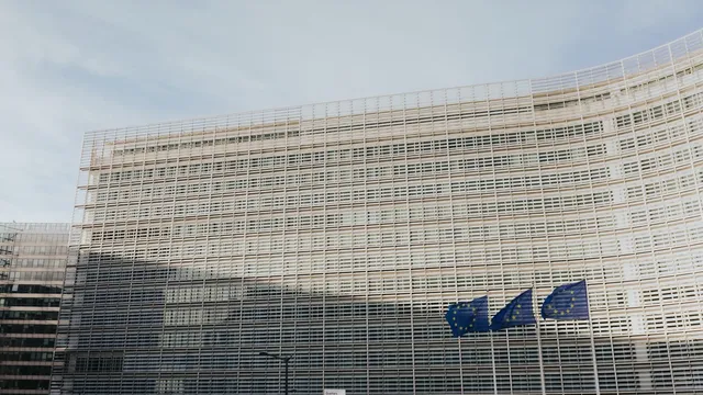 The Berlaymont building in Brussels, headquarters of the European Commission, its cross-shaped glass facade seen from the street with European flags in front.