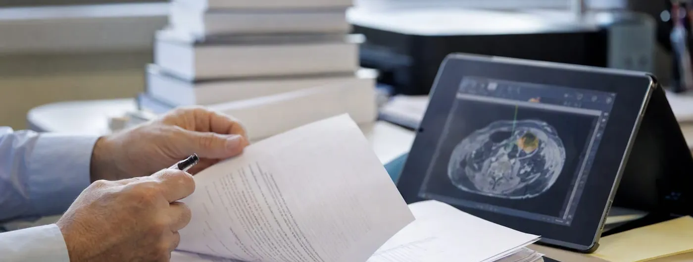 Photorealistic close-up of a pair of hands turning printed policy documents beside a tablet displaying a medical imaging scan with a software-generated overlay, on a desk in an office.