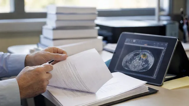 Photorealistic close-up of a pair of hands turning printed policy documents beside a tablet displaying a medical imaging scan with a software-generated overlay, on a desk in an office.