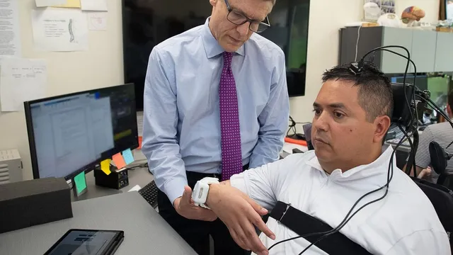 In a laboratory, Chad Bouton steadies the forearm of participant Keith Thomas, who sits in a wheelchair with cabling attached to his head and a white wearable stimulation patch on his wrist.