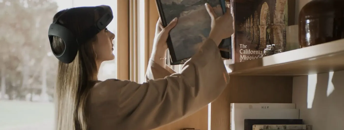 A woman wearing a dark head-mounted recording rig holds a large book up to a shelf on a wooden bookcase in a sunlit living room, her face in profile beside a tall window.