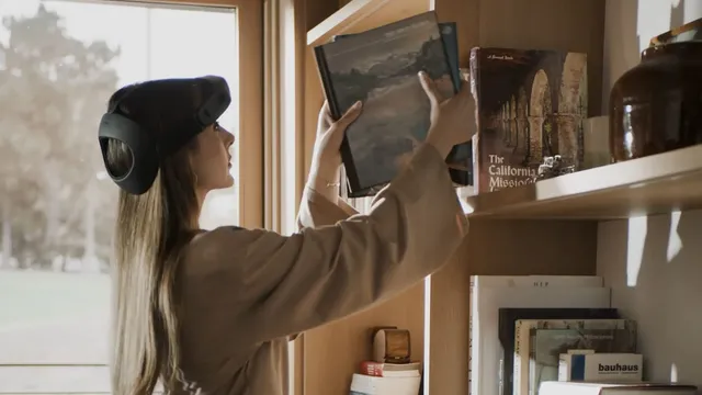 A woman wearing a dark head-mounted recording rig holds a large book up to a shelf on a wooden bookcase in a sunlit living room, her face in profile beside a tall window.
