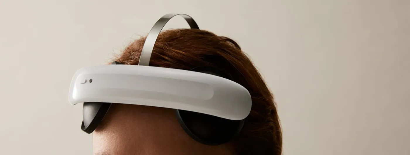 A woman wears the white and black Flow Neuroscience FL-100 headset, its curved band resting across her forehead above the eyes with a slim metal strap running over the top of her head.