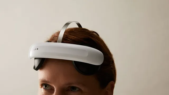 A woman wears the white and black Flow Neuroscience FL-100 headset, its curved band resting across her forehead above the eyes with a slim metal strap running over the top of her head.