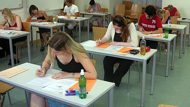 University students seated at desks in a large hall, writing an examination on paper.