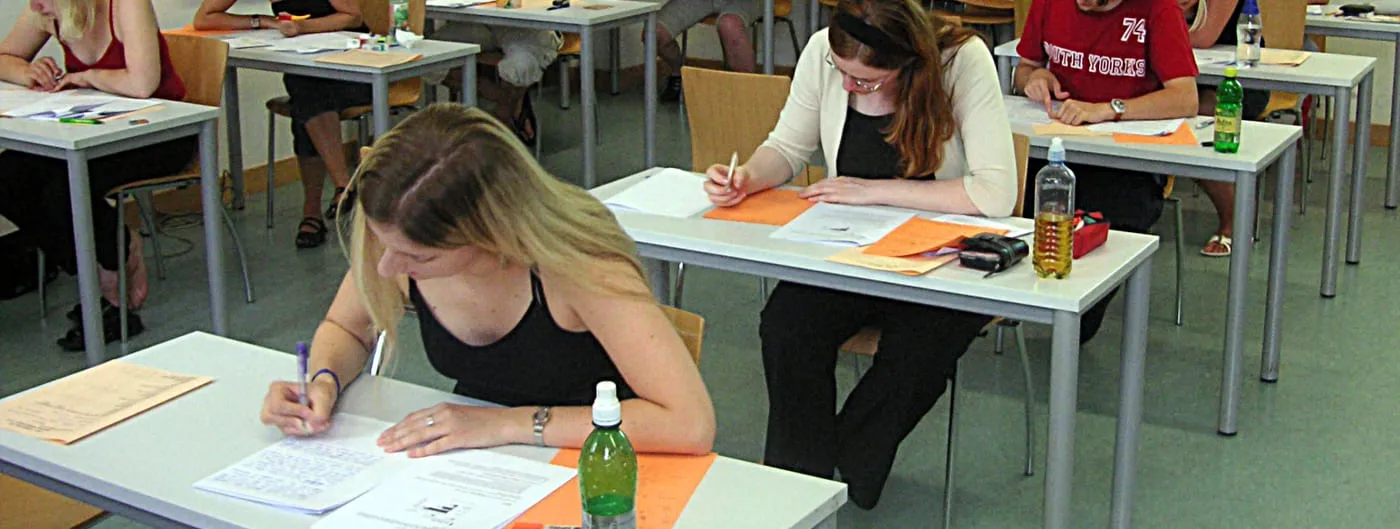 University students seated at desks in a large hall, writing an examination on paper.