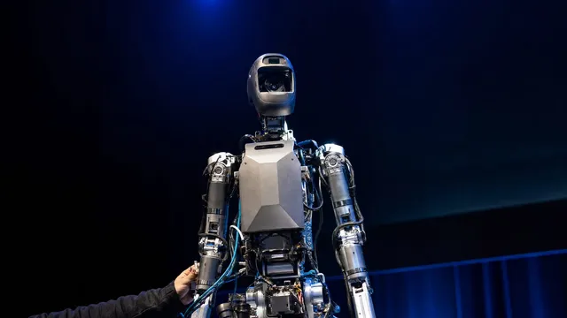 Photorealistic waist-up shot of a bipedal humanoid robot's head and torso on an exhibition stage under blue stage lighting, with generous headroom above and an engineer's hand adjusting cables at its base.
