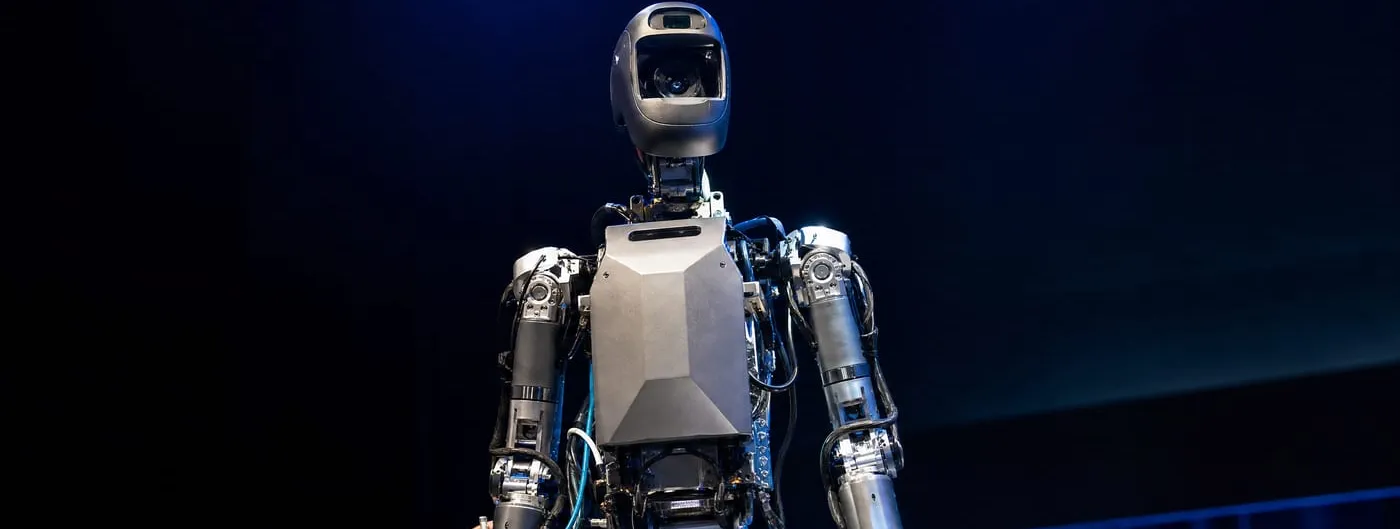 Photorealistic waist-up shot of a bipedal humanoid robot's head and torso on an exhibition stage under blue stage lighting, with generous headroom above and an engineer's hand adjusting cables at its base.