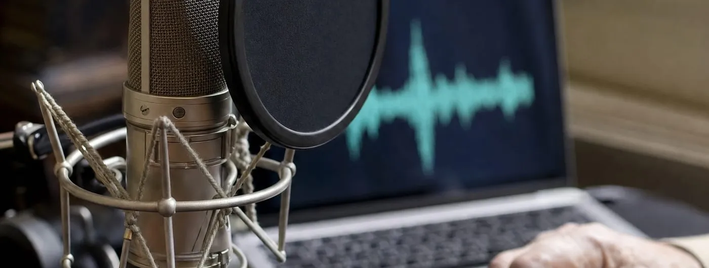 A studio condenser microphone with a pop filter in the foreground, a laptop behind it showing a blurred teal audio waveform, and an older person hand resting on the desk.