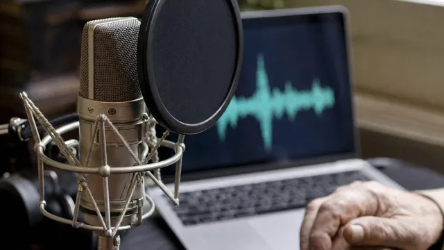 A studio condenser microphone with a pop filter in the foreground, a laptop behind it showing a blurred teal audio waveform, and an older person hand resting on the desk.