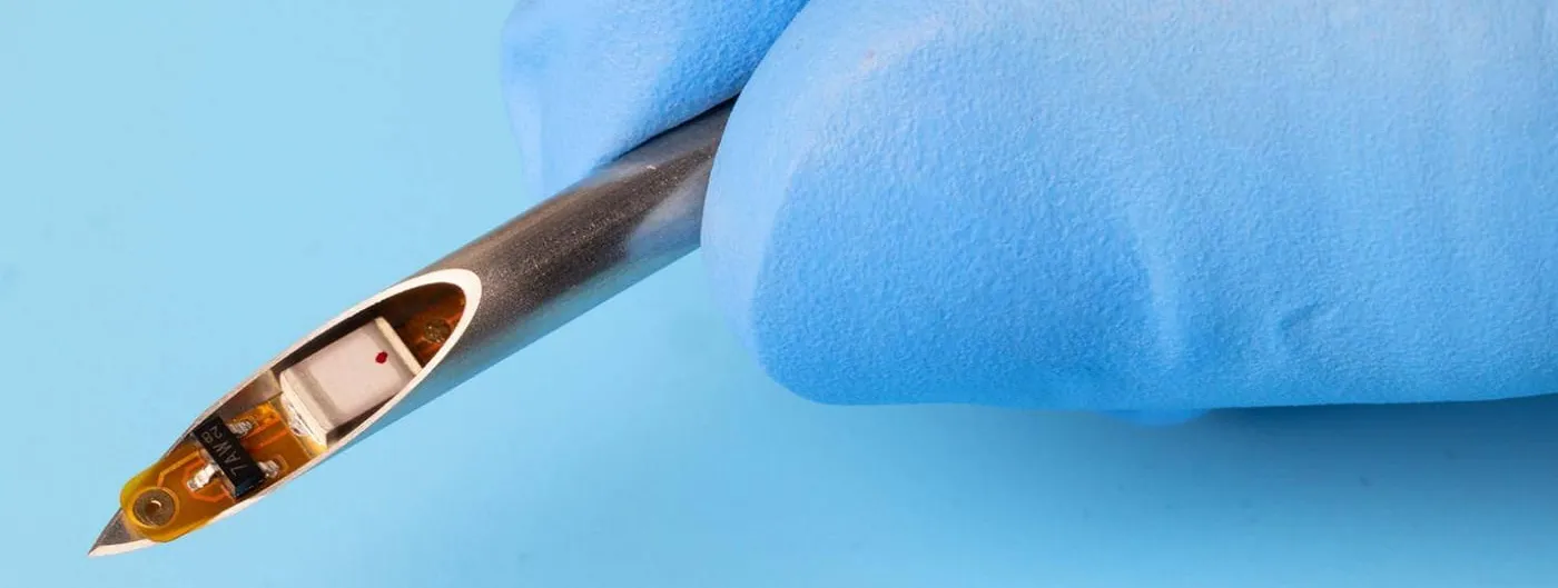 A gloved hand holds a syringe needle whose open bevel reveals a tiny implant, a small circuit on a flexible amber substrate beside a white component, against a light blue background.