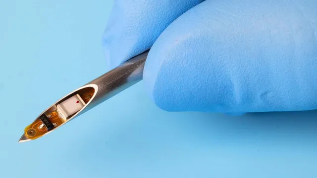 A gloved hand holds a syringe needle whose open bevel reveals a tiny implant, a small circuit on a flexible amber substrate beside a white component, against a light blue background.