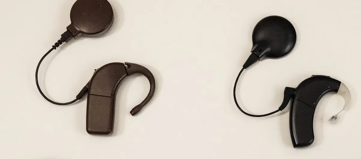 Two behind-the-ear cochlear implant sound processors, one brown and one black, each trailing a thin cable to a round magnetic transmitter coil, laid out on a plain cream background.