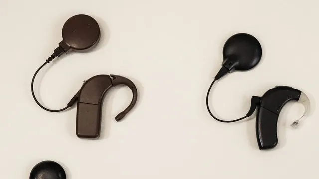Two behind-the-ear cochlear implant sound processors, one brown and one black, each trailing a thin cable to a round magnetic transmitter coil, laid out on a plain cream background.