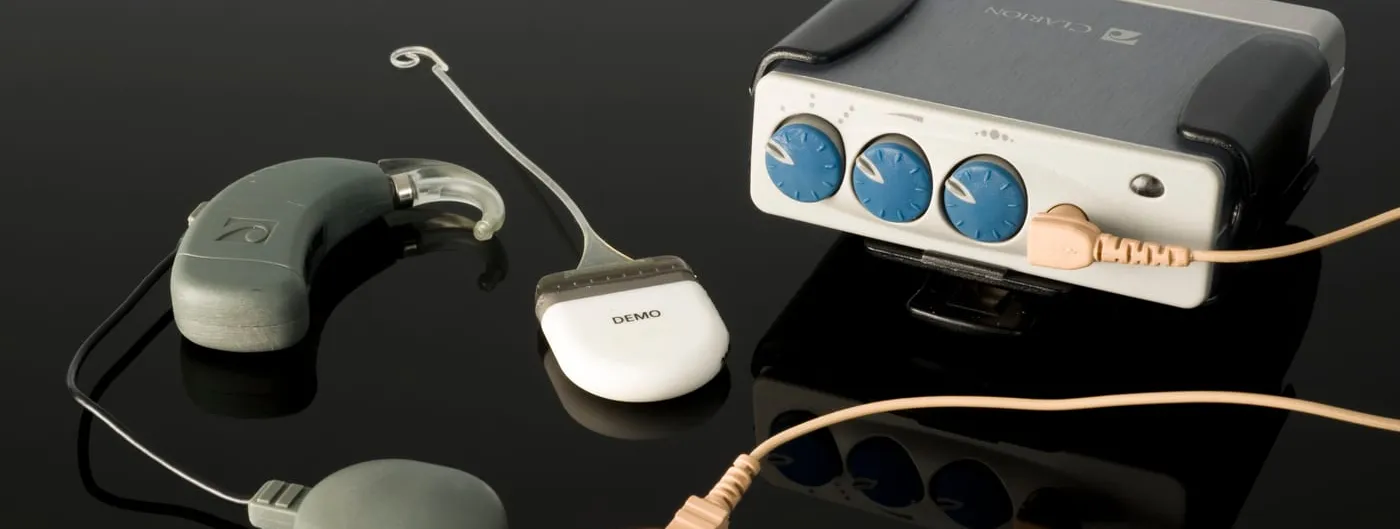 A cochlear implant kit laid out on a reflective black surface, showing the behind-the-ear headpiece, a transmitting coil with a magnet, and an external speech processor with three tuning dials, connected by beige cables.