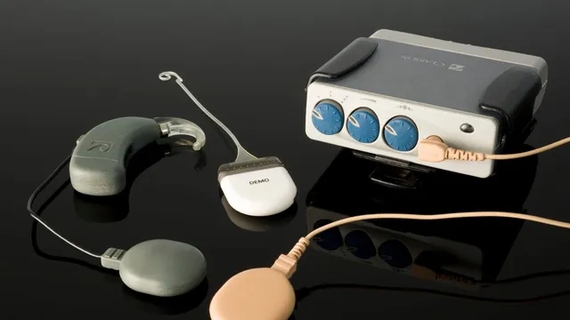 A cochlear implant kit laid out on a reflective black surface, showing the behind-the-ear headpiece, a transmitting coil with a magnet, and an external speech processor with three tuning dials, connected by beige cables.