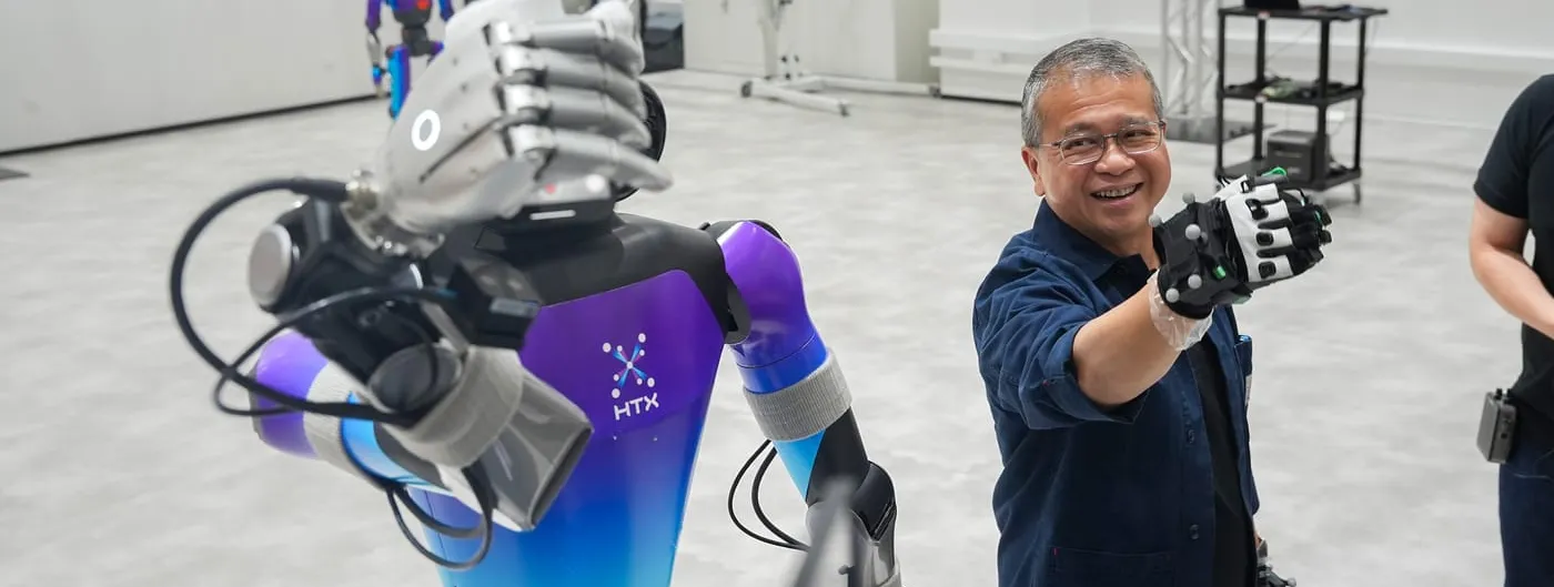 A humanoid robot with a purple-and-blue torso marked HTX raises its articulated hand toward the camera inside a bright test hall, while a smiling man in a dark blue shirt wearing motion-capture gloves mirrors the gesture beside it.