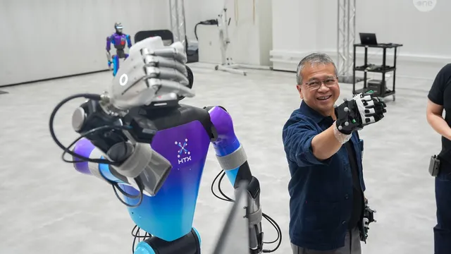 A humanoid robot with a purple-and-blue torso marked HTX raises its articulated hand toward the camera inside a bright test hall, while a smiling man in a dark blue shirt wearing motion-capture gloves mirrors the gesture beside it.