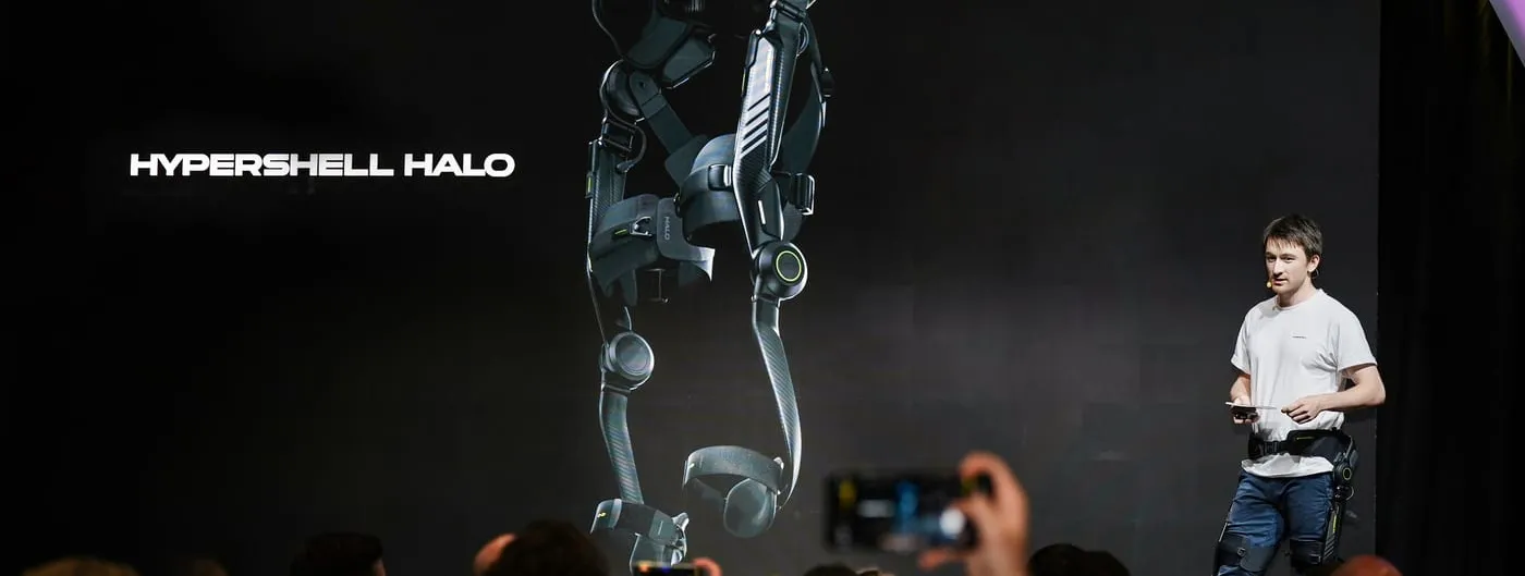 A presenter in a white shirt stands on a dark stage beside a large screen showing a full-leg exoskeleton with hip and knee joints, while seated attendees raise phones to photograph it.