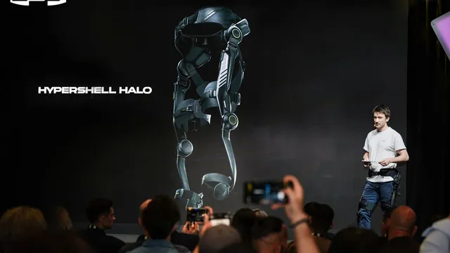 A presenter in a white shirt stands on a dark stage beside a large screen showing a full-leg exoskeleton with hip and knee joints, while seated attendees raise phones to photograph it.