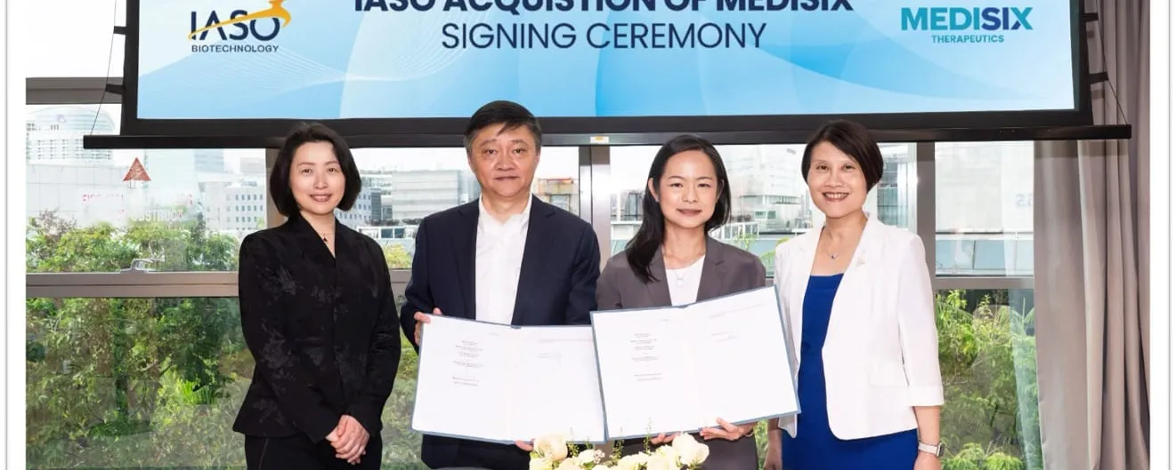 Four executives stand in a glass-walled meeting room holding open signed folders toward the camera, beneath a screen reading IASO Acquisition of MediSix Signing Ceremony with both company logos.