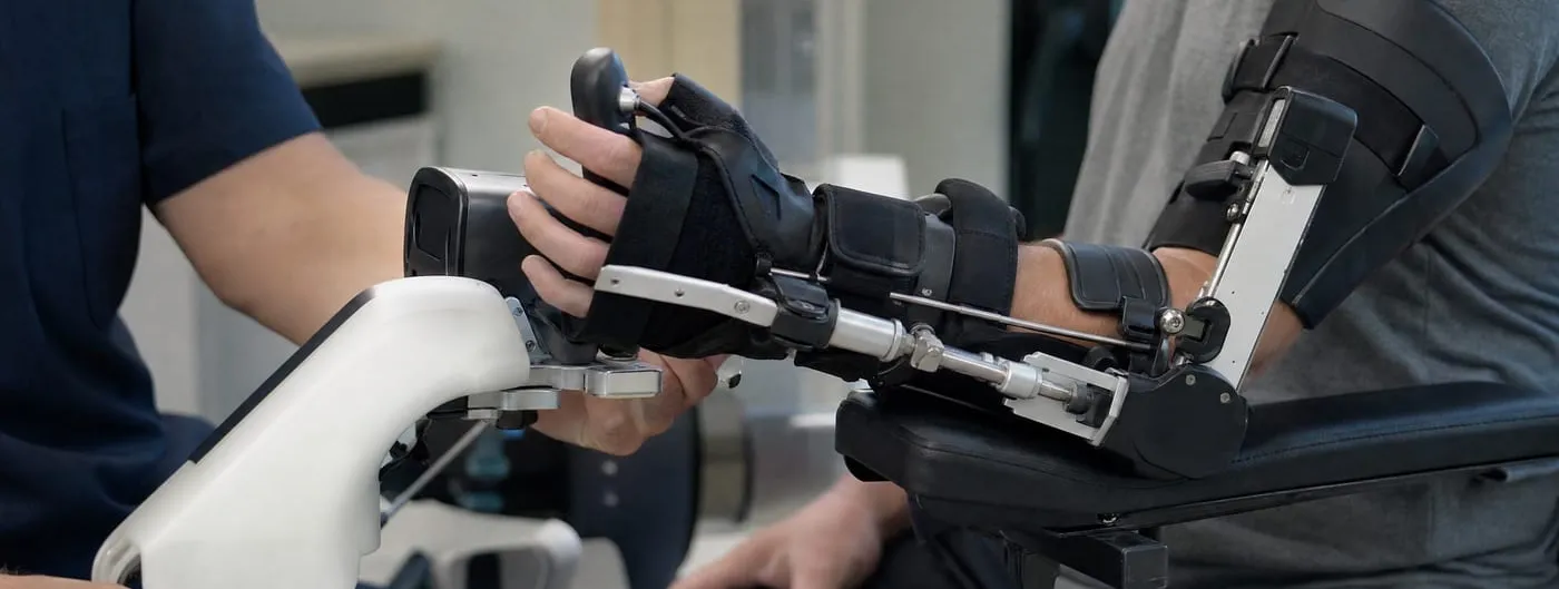 A motorised hand and forearm rehabilitation exoskeleton strapped to the arm of a seated person in a clinic, with a therapist supporting the device frame by hand.