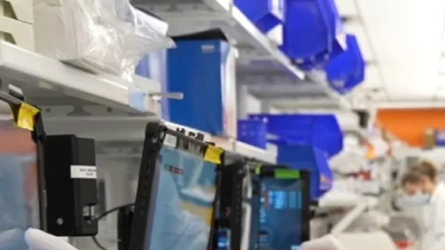 A laboratory interior at Intellia Therapeutics, with instrument touchscreens in the foreground, shelves of blue-lidded containers overhead, and a masked worker in a white coat at the back of the room.