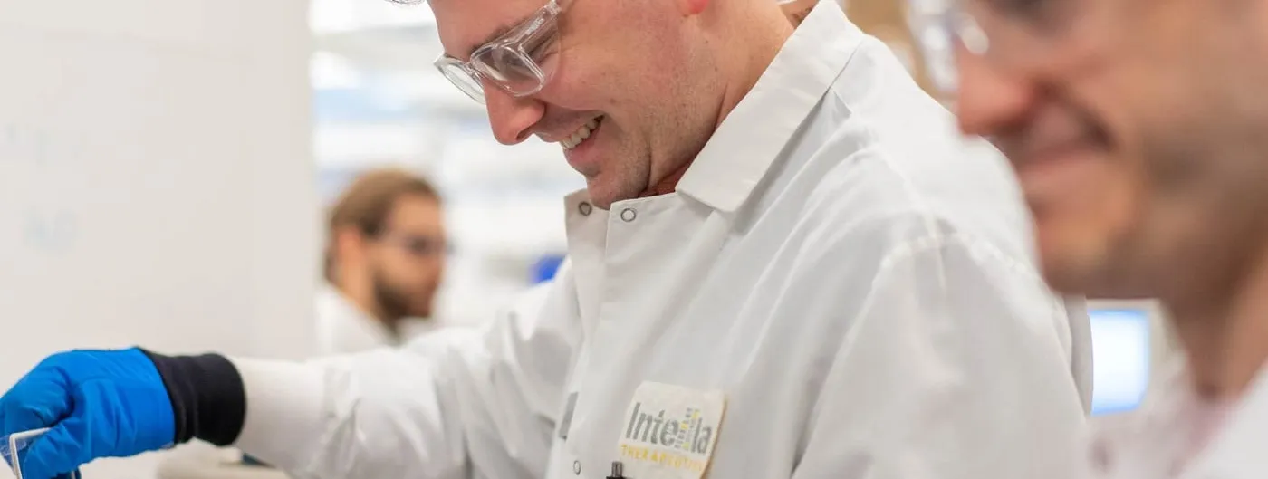 A scientist in safety glasses and a white coat badged Intellia Therapeutics reaches into a metal cryogenic container on a laboratory bench, with colleagues working out of focus behind.