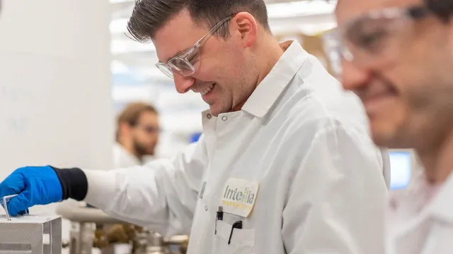 A scientist in safety glasses and a white coat badged Intellia Therapeutics reaches into a metal cryogenic container on a laboratory bench, with colleagues working out of focus behind.