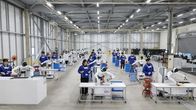 Rows of desks in a bright industrial hall, each with a small humanoid robot arm rig, attended by operators in blue uniforms.