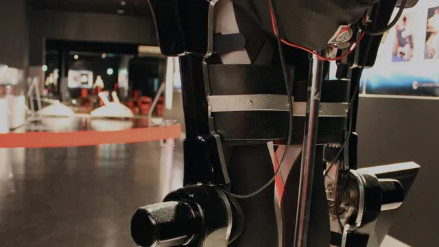 Close-up of a HAL powered exoskeleton's leg unit on museum display, showing its black actuator housings, metal knee and ankle joints, straps and wiring against a blurred gallery background.