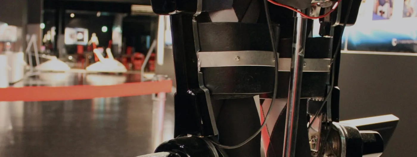 Close-up of a HAL powered exoskeleton's leg unit on museum display, showing its black actuator housings, metal knee and ankle joints, straps and wiring against a blurred gallery background.