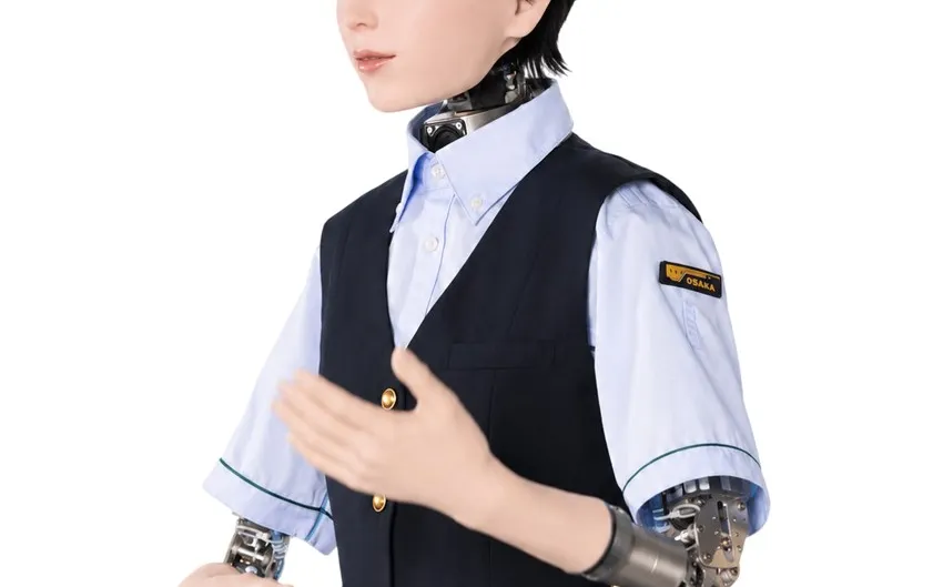An android in a navy Osaka Metro waistcoat, pale blue shirt and peaked cap gestures with one hand, its exposed metal wrist and forearm mechanism visible below the rolled sleeve.
