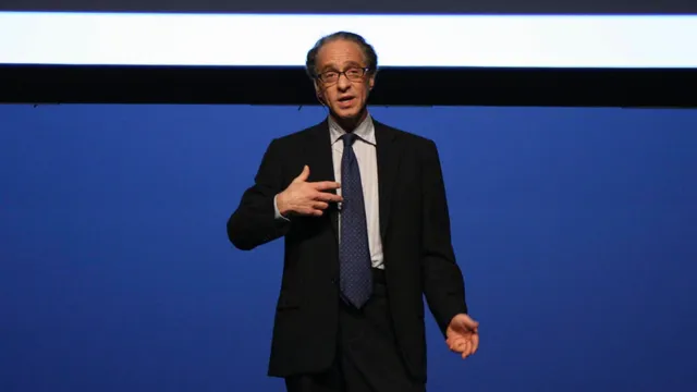 Ray Kurzweil, in a dark suit and patterned tie, speaks with one hand raised while standing on a stage against a plain deep-blue backdrop.