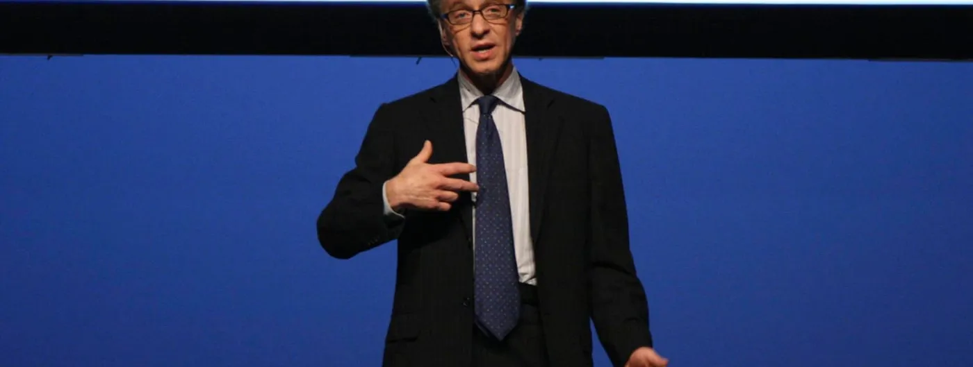 Ray Kurzweil, in a dark suit and patterned tie, speaks with one hand raised while standing on a stage against a plain deep-blue backdrop.