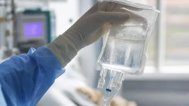 A gloved clinician's hands hold up a clear infusion bag of cell-therapy fluid beside a hospital bed, with a monitor screen visible in the background.
