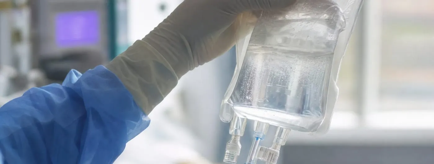 A gloved clinician's hands hold up a clear infusion bag of cell-therapy fluid beside a hospital bed, with a monitor screen visible in the background.