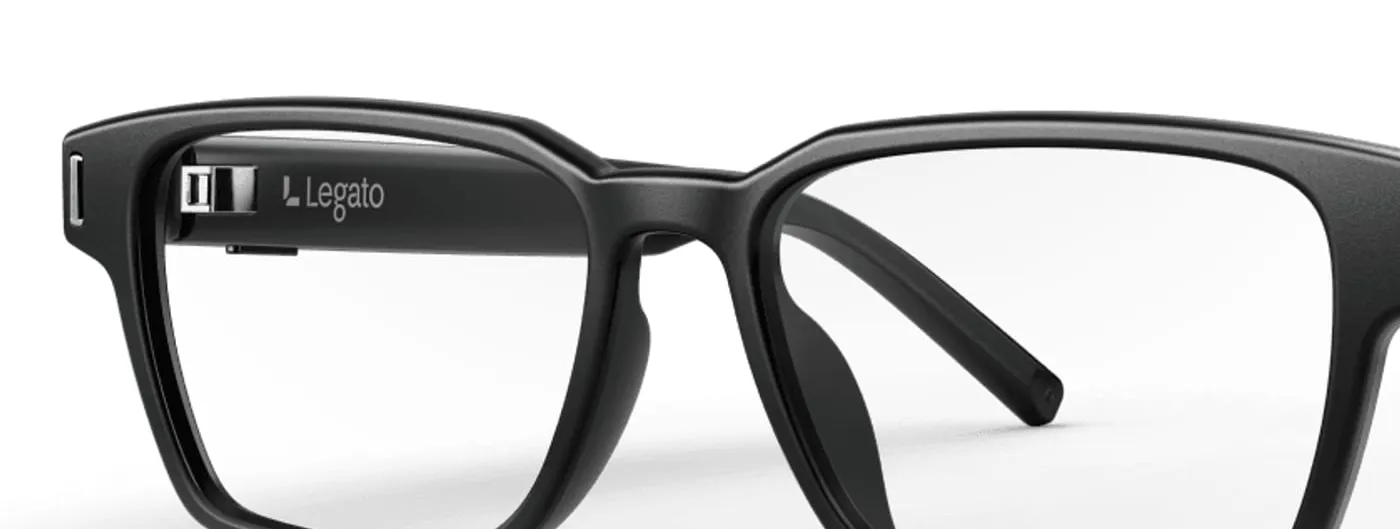 A close-up of matte black eyeglass frames on a white background, with a small module and the Legato name visible inside the left temple arm.