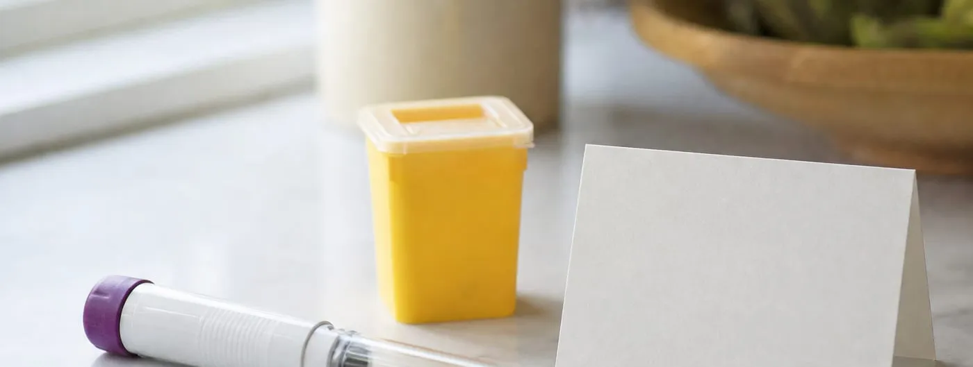 A subcutaneous autoinjector pen lies on a pale kitchen counter beside a small yellow sharps container and a blank folded card, lit by a nearby window.