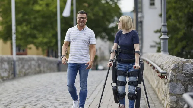 A woman wearing a ReWalk lower-limb exoskeleton, its leg braces and hip motor visible, walks upright using forearm crutches across a cobblestone bridge beside a companion.