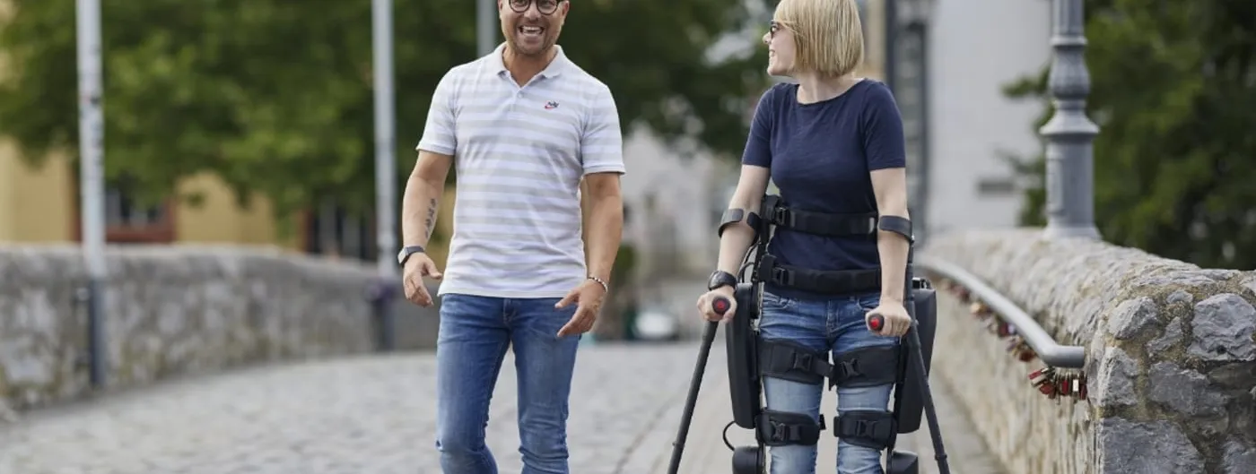 A woman wearing a ReWalk lower-limb exoskeleton, its leg braces and hip motor visible, walks upright using forearm crutches across a cobblestone bridge beside a companion.