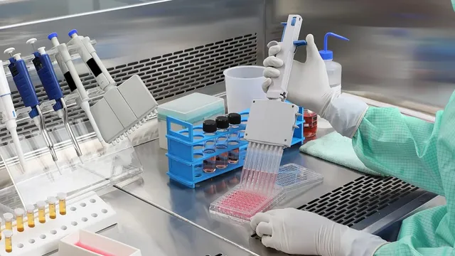 A gloved lab technician in a cleanroom gown handles a multichannel pipette over a 96-well plate of pink cell-culture media on a stainless steel bench.