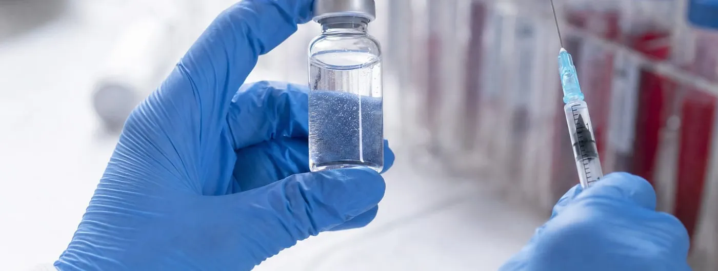 A gloved researcher holds a small glass vial of clear liquid beside a syringe on a laboratory bench, with racks of sample tubes blurred in the background.
